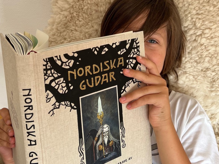 Barn sitter i fåtölj och kikar fram bakom en bok om nordiska gudar
